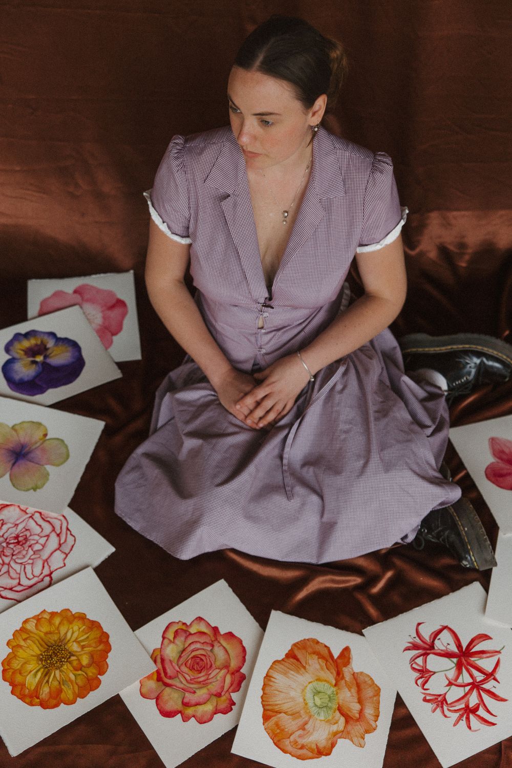 Artist in purple dress arranges colorful floral watercolor paintings on brown fabric.