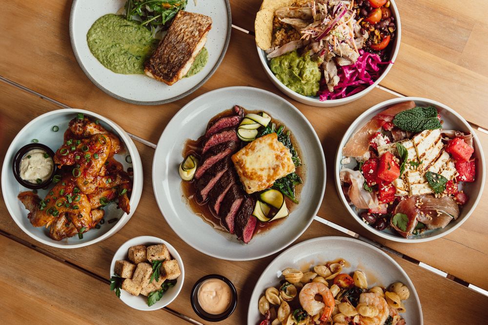 Flatlay of diverse restaurant dishes including seafood, meats, and sides on white plates.