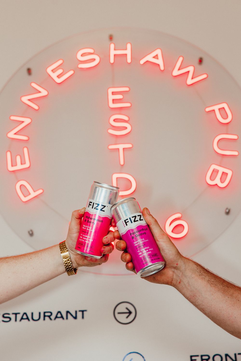 Pink neon sign and hands holding canned beverages in a restaurant setting.