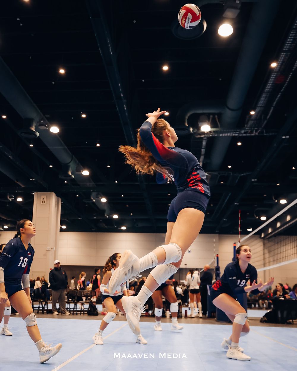 A female volleyball player leaps to spike the ball during an indoor tournament game, teammates visible in background.