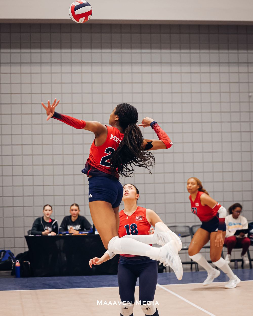 Volleyball player in red jersey jumping to spike the ball during an indoor match, teammates visible in background.