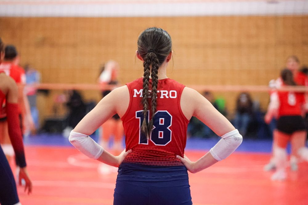 Female volleyball player wearing red Metro jersey number 18 stands with hands on hips during indoor tournament.