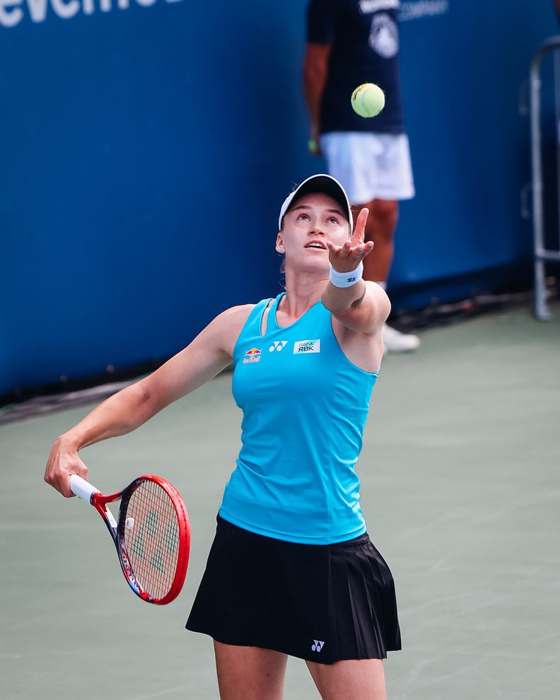 Female tennis player in teal Yonex outfit serving ball on outdoor court during competitive match.