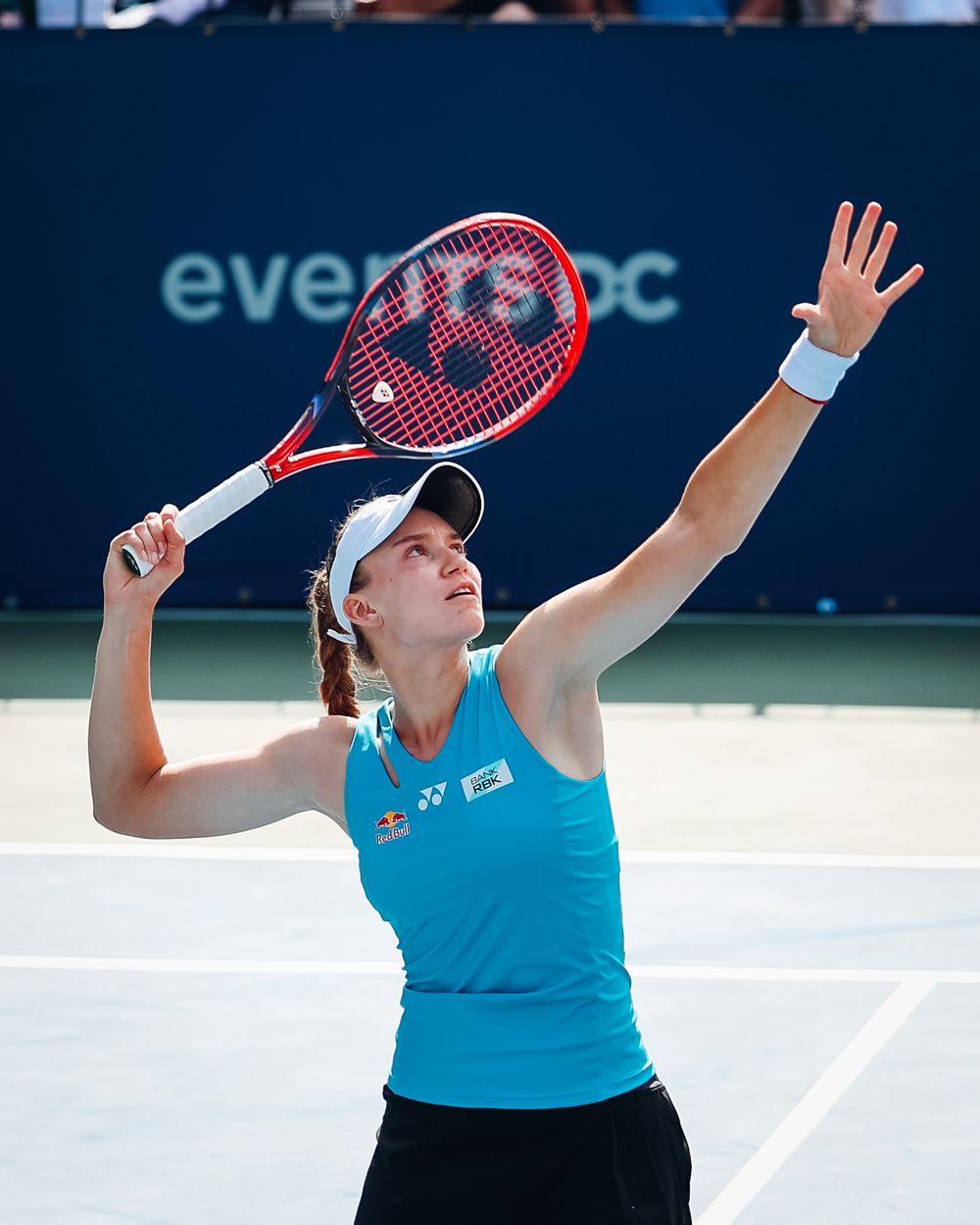 Female tennis player in blue top serving with a Yonex racket on an outdoor hard court during a professional match.