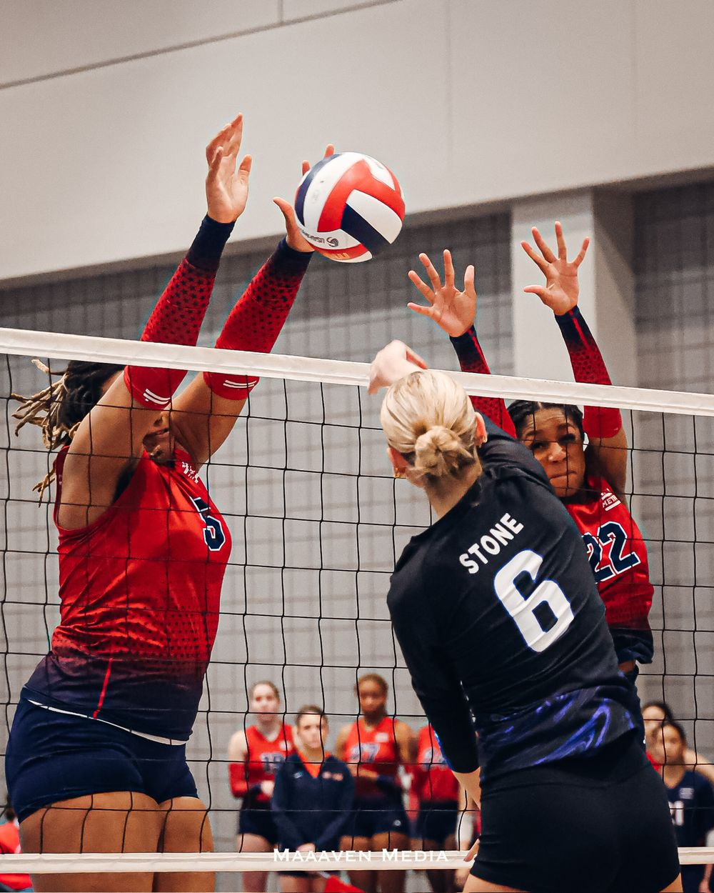 Two volleyball players in red block a spike attempt by player Stone #6 in a competitive indoor volleyball match.