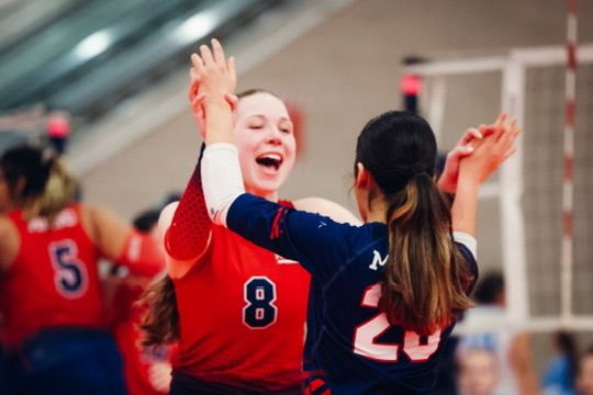 Two volleyball players in red and blue uniforms celebrate with a high-five during an indoor match.