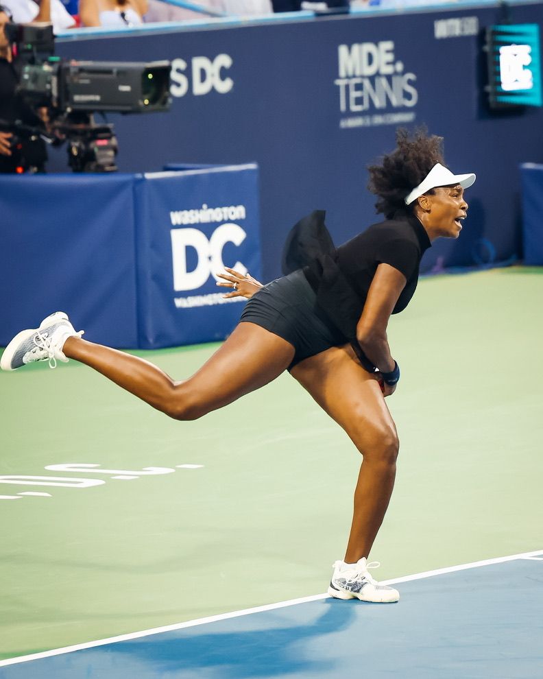 Female tennis player mid-serve on court at Washington DC MDE Tennis tournament, wearing black outfit and white shoes.
