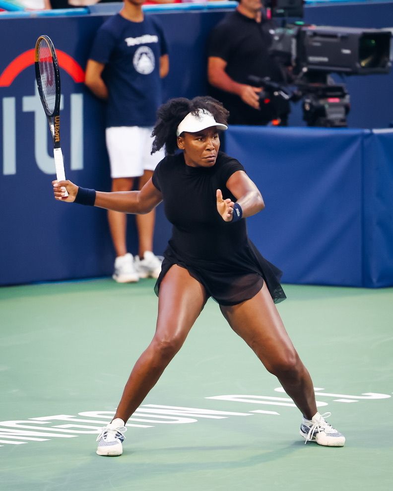Female tennis player in black outfit mid-stance on court, holding racket ready to return shot.