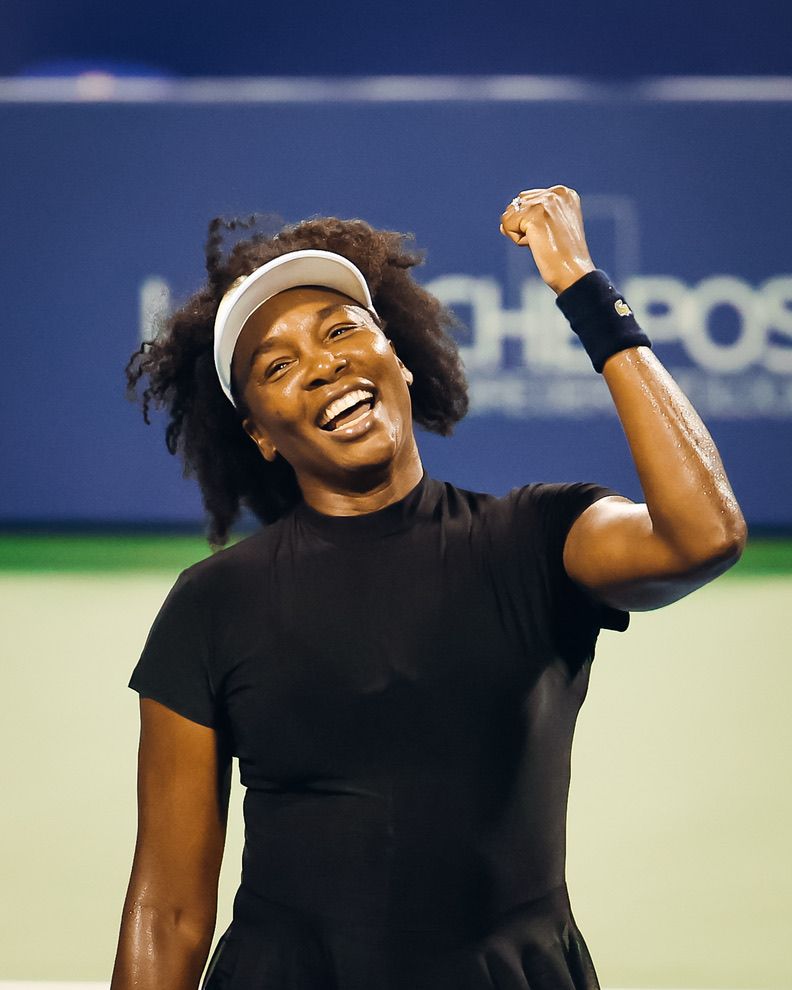 Female tennis player in black outfit celebrates victory with raised fist and joyful smile on court.