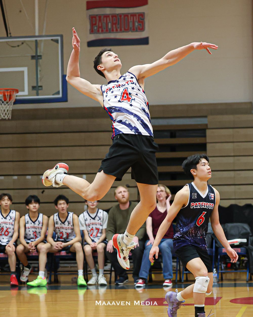 A Patriots basketball player jumps to shoot during an indoor high school game while teammates watch from the bench.