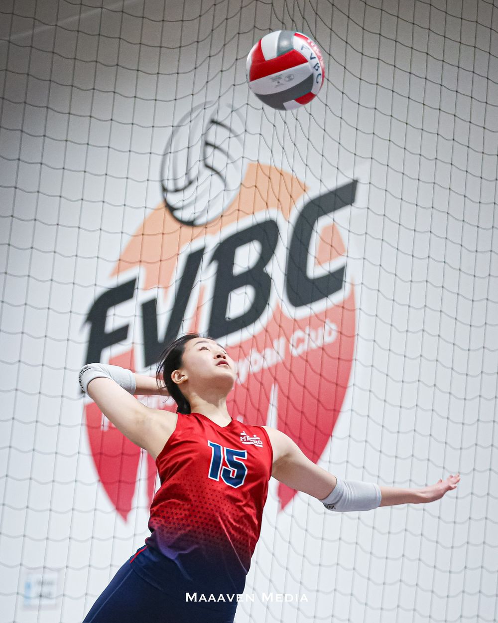 Female volleyball player #15 in red jersey jumps to spike ball at FVBC Volleyball Club indoor court.