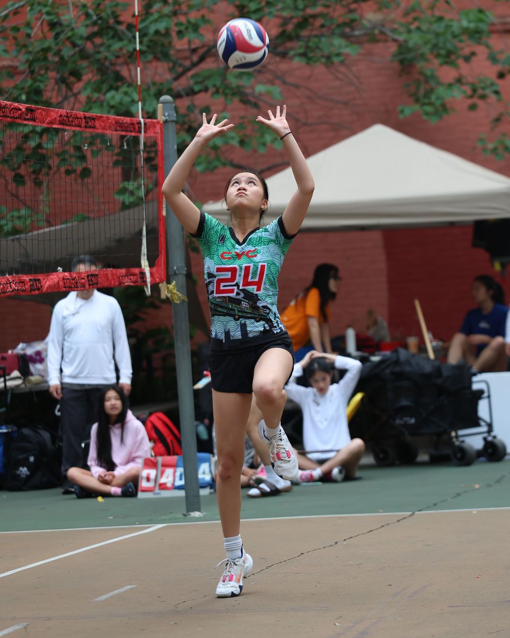 Player #24 in green jersey leaps to set a volleyball during an outdoor netball match.