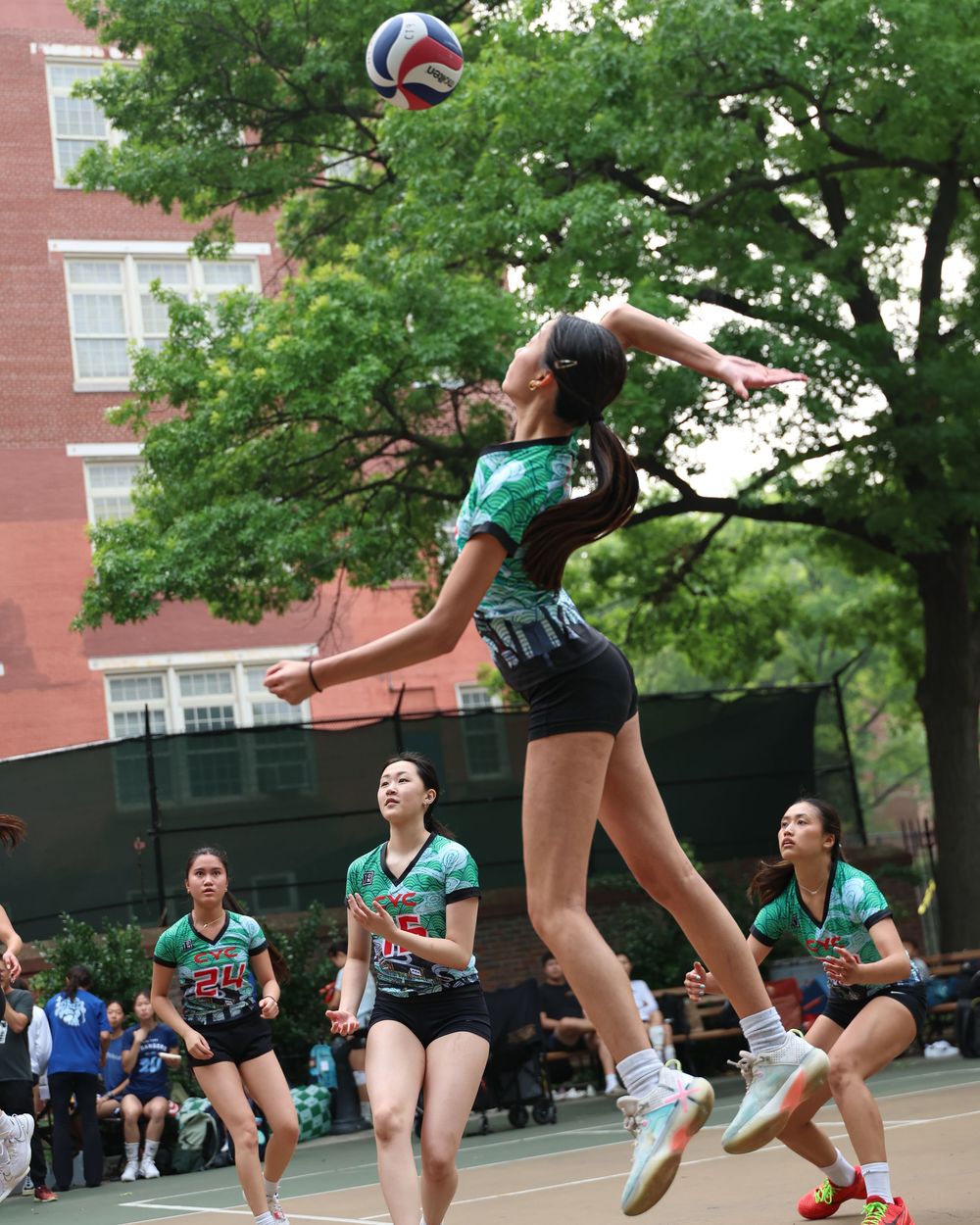 Female volleyball player jumps to spike ball during outdoor court game, teammates ready in green uniforms.