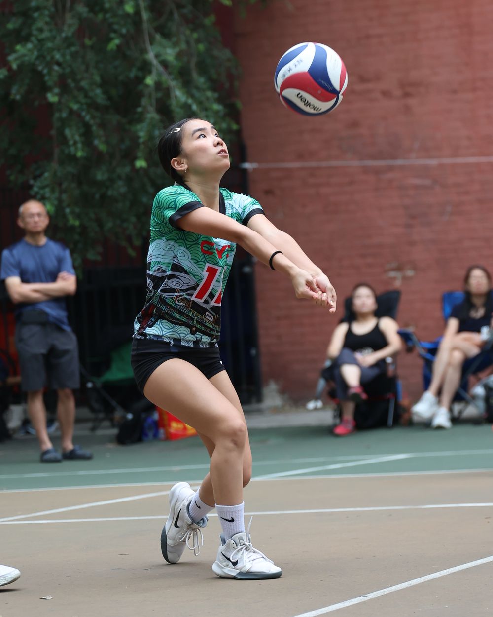 A young female volleyball player in a green jersey bumps the ball during an outdoor court game.
