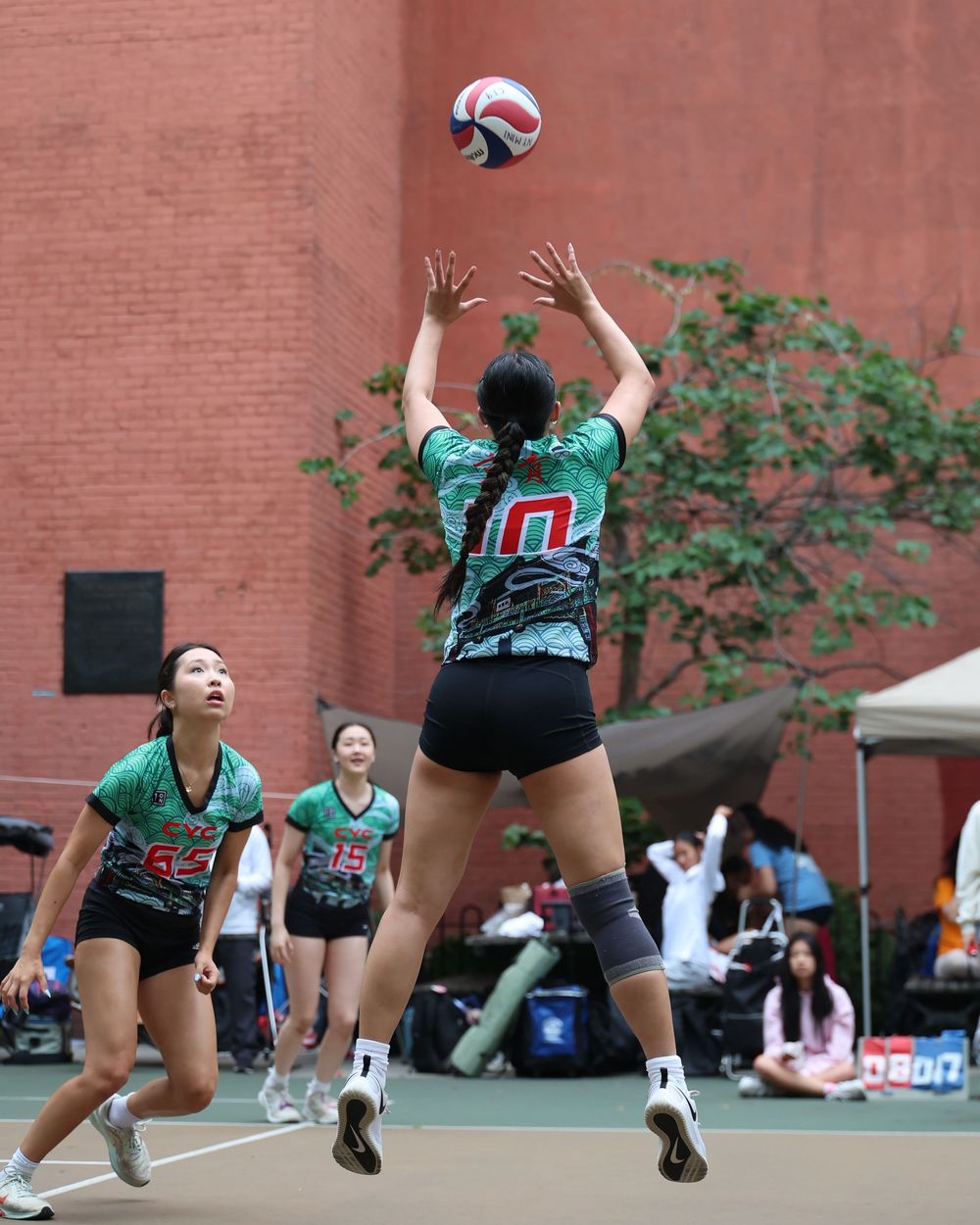 Female volleyball players in green jerseys compete outdoors, with player #10 setting the ball during a match near a brick wall.