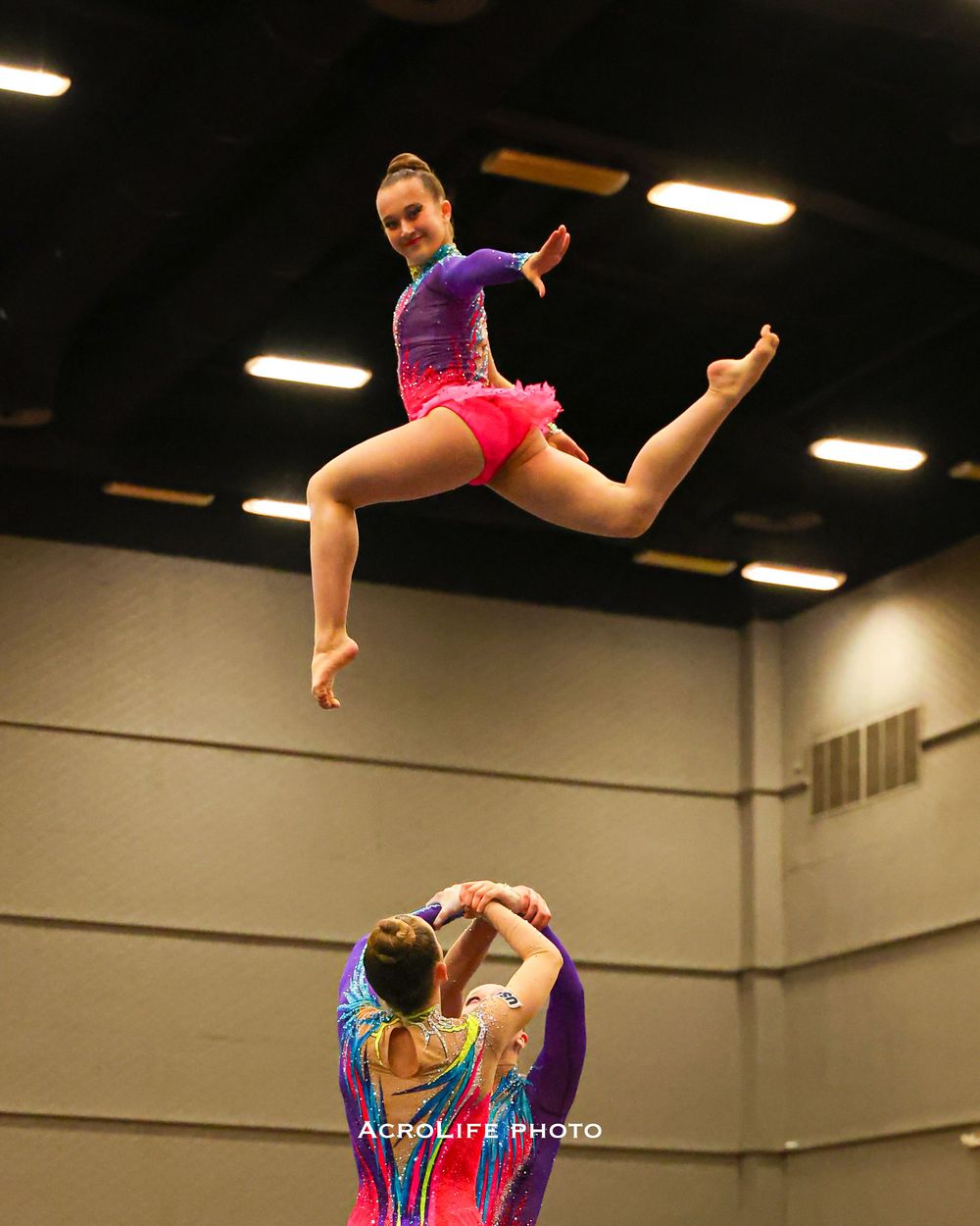 Young acrobatic gymnast leaping mid-air in purple leotard while partner supports below during competition.