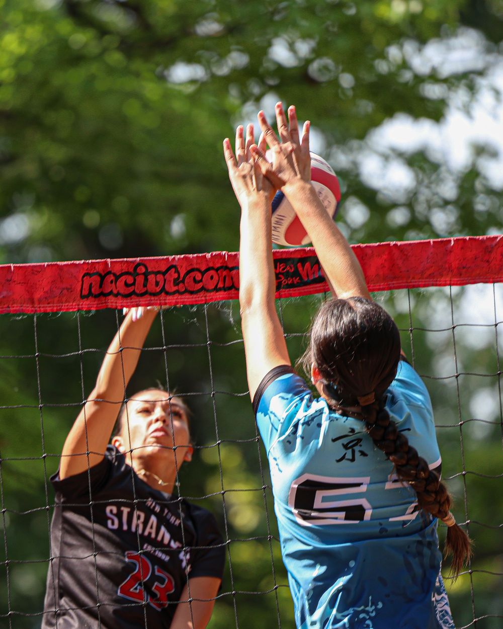 Two volleyball players battle at the net during an outdoor match, one in black jersey number 23, the other in blue number 5.