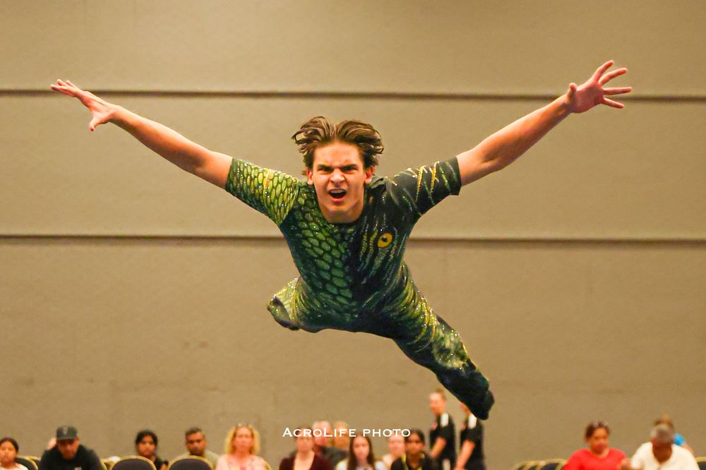 Acrobat in green costume leaps with arms spread wide at an indoor performance event.