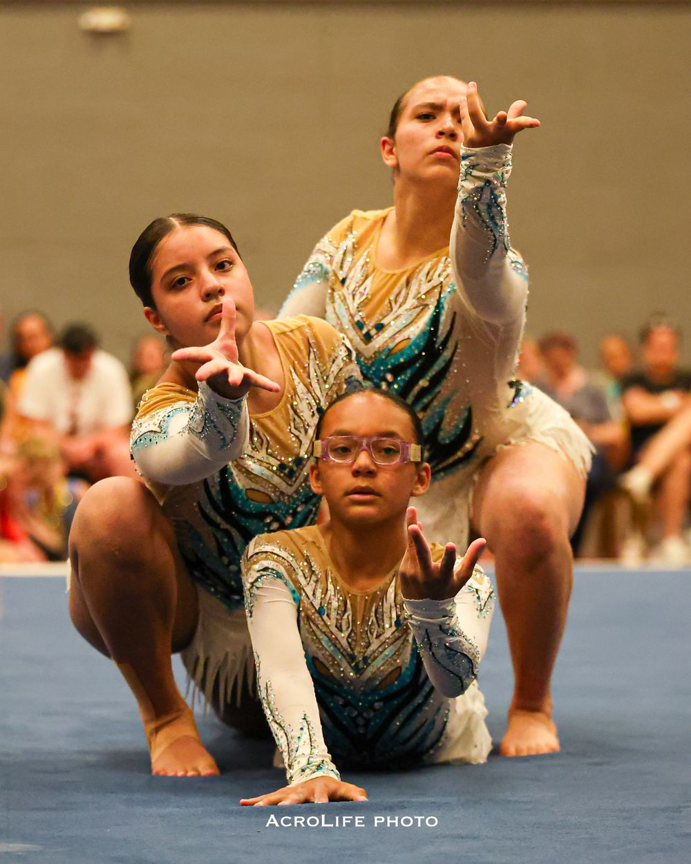 Three acrobatic gymnasts in matching teal and gold sequined leotards pose dramatically on the floor during a competition.