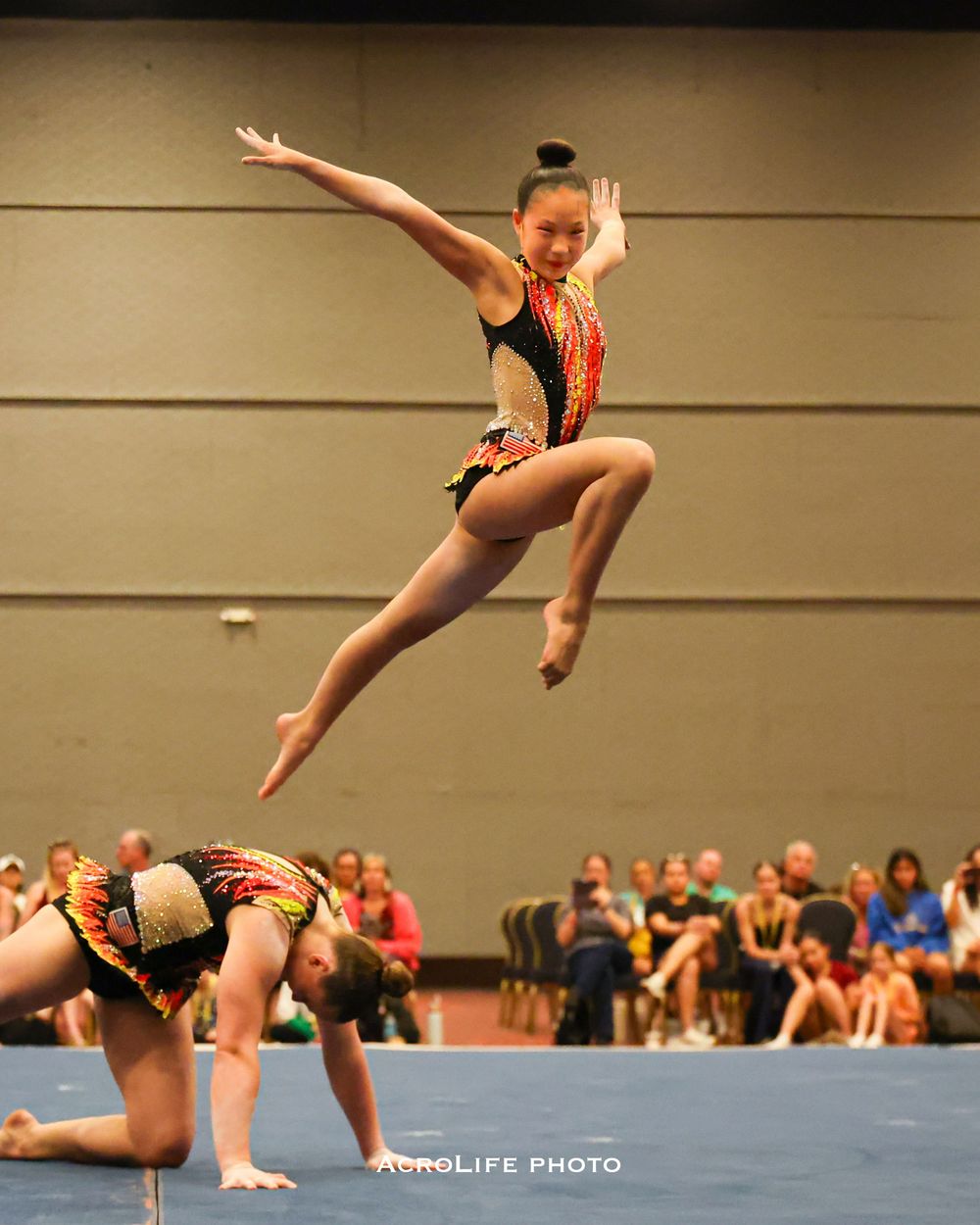 Gymnast in red and black leotard leaps high above teammates during acrobatic gymnastics competition.