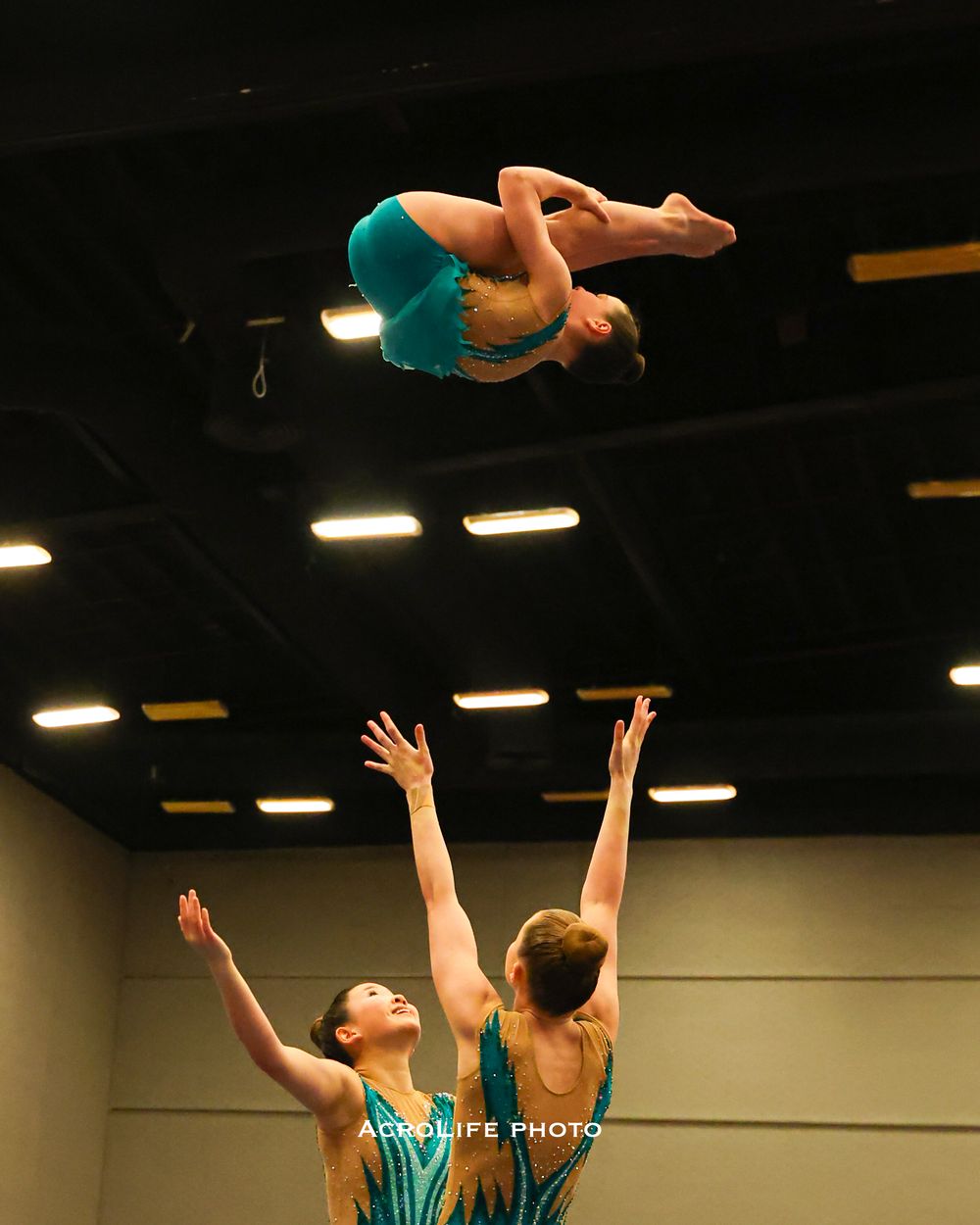 Three acrobatic gymnasts performing a throw, with one athlete soaring high in teal leotard.