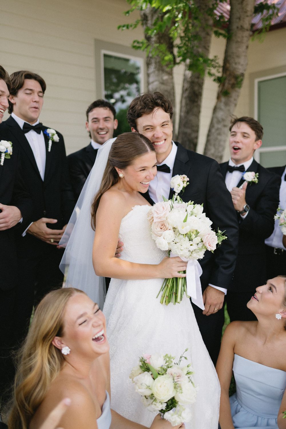Bride in strapless white gown holds bouquet surrounded by cheering groomsmen in black tuxedos.