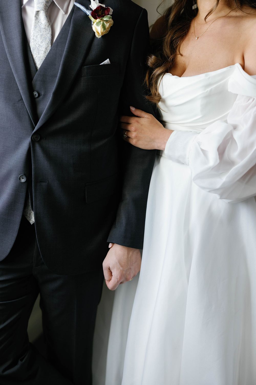 Close-up of bride in white off-shoulder gown holding grooms hand as she leans against his dark suit.