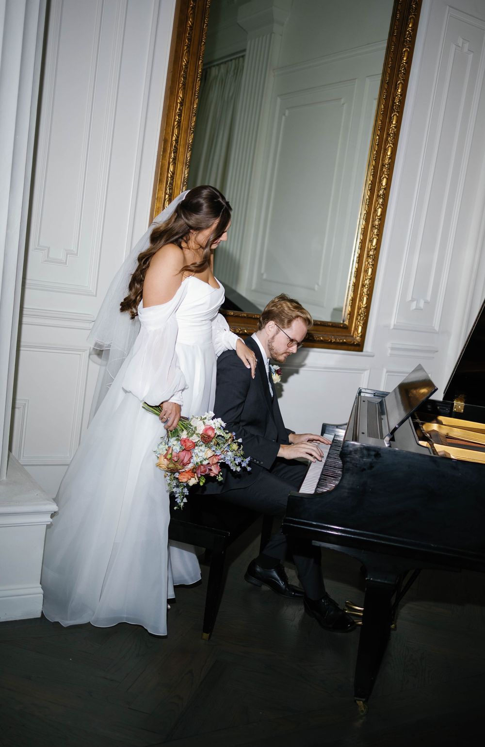 Bride holding floral bouquet stands beside groom playing grand piano before ornate gold mirror.