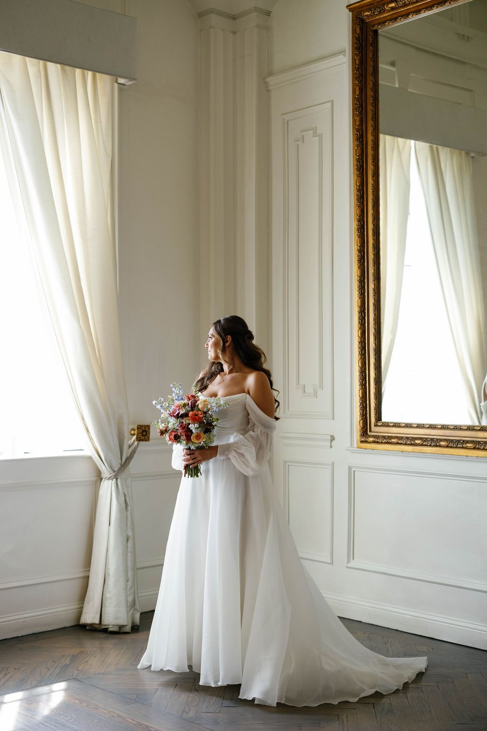Elegant bride in off-shoulder white gown holds colorful bouquet while gazing out window by gold mirror.