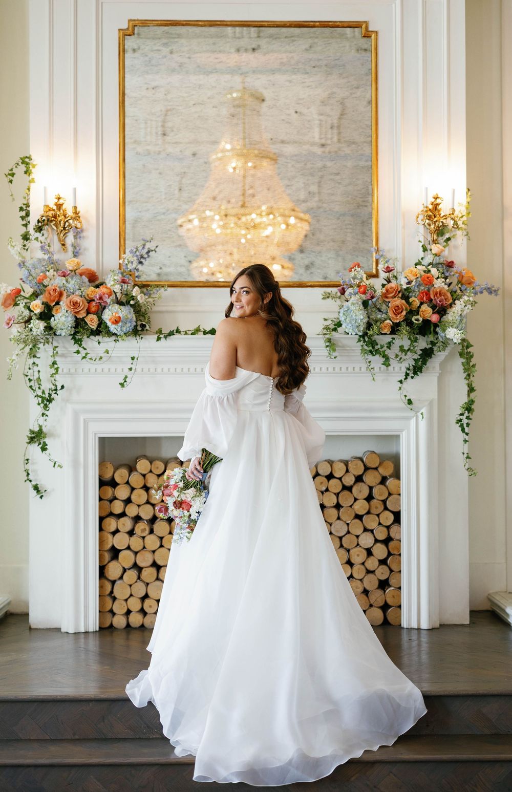 Bride in flowing off-shoulder white gown poses before ornate fireplace adorned with colorful floral arrangements.