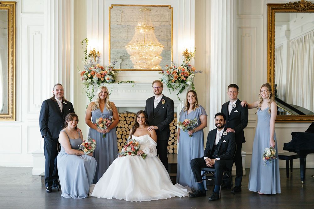 Wedding party poses in elegant white ballroom with chandelier, fireplace florals, and grand piano.