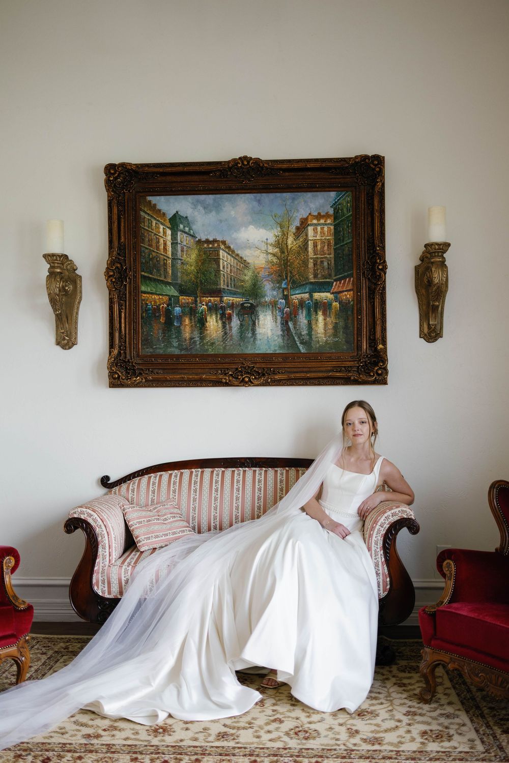 Bride in a white gown with long veil seated on an antique sofa beneath a framed painting.