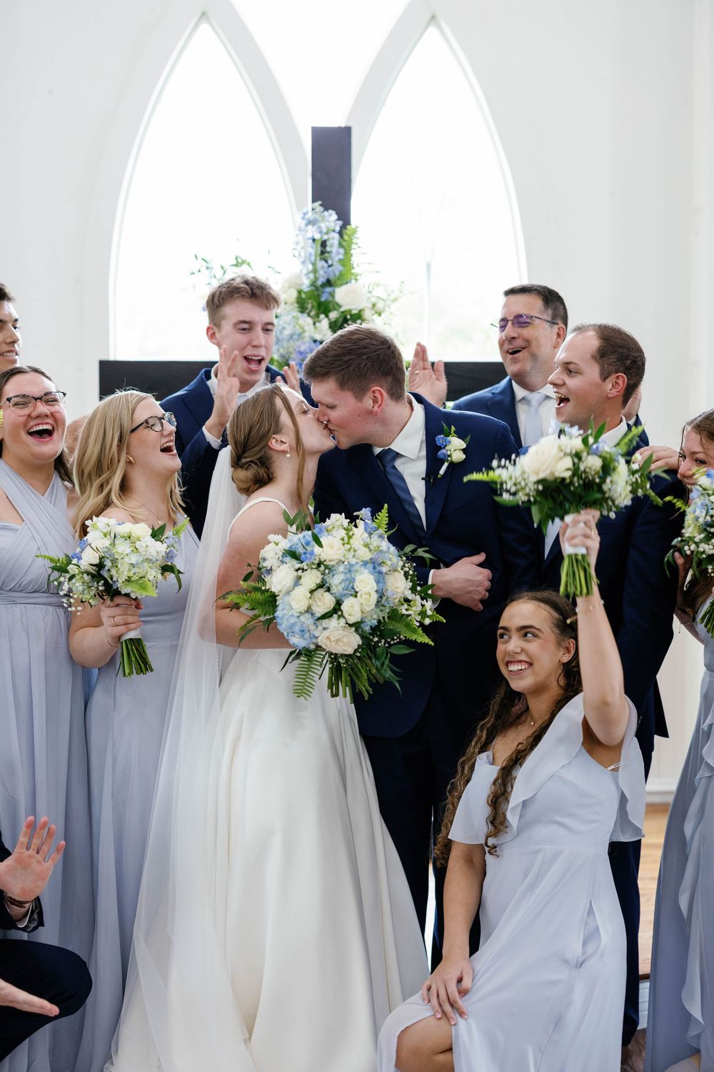 Bride and groom kissing at the altar surrounded by cheering wedding party in a white chapel.