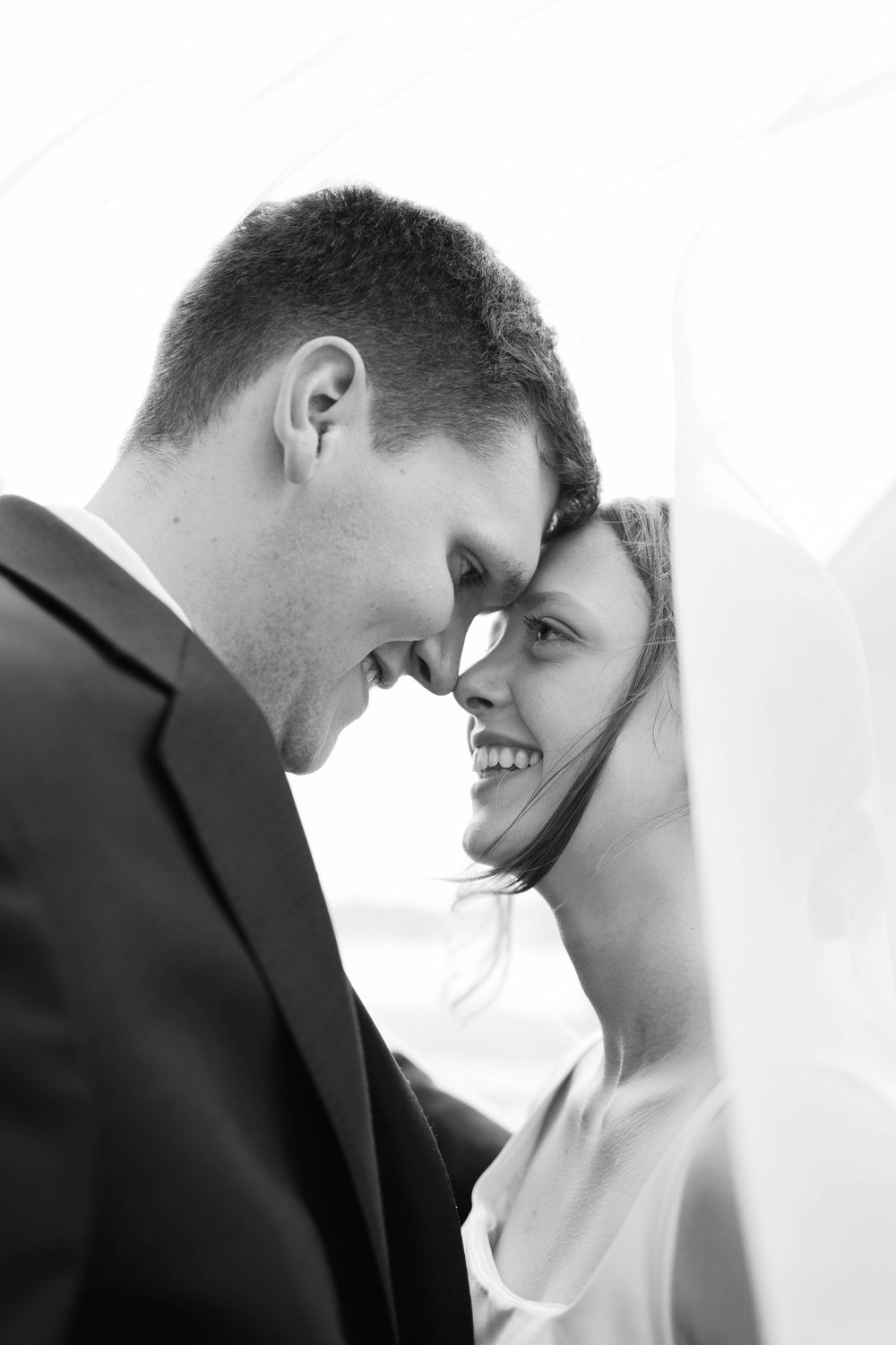 Black and white close-up of smiling bride and groom touching foreheads romantically under a flowing veil.