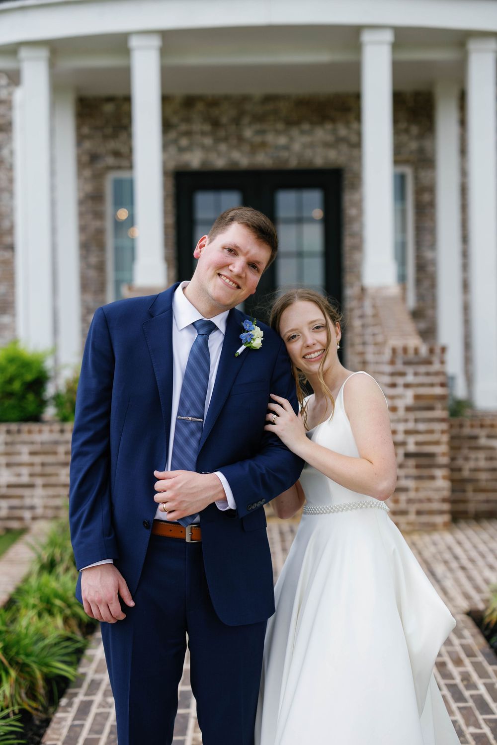 Smiling bride and groom pose together outside elegant brick mansion with white columns on wedding day.