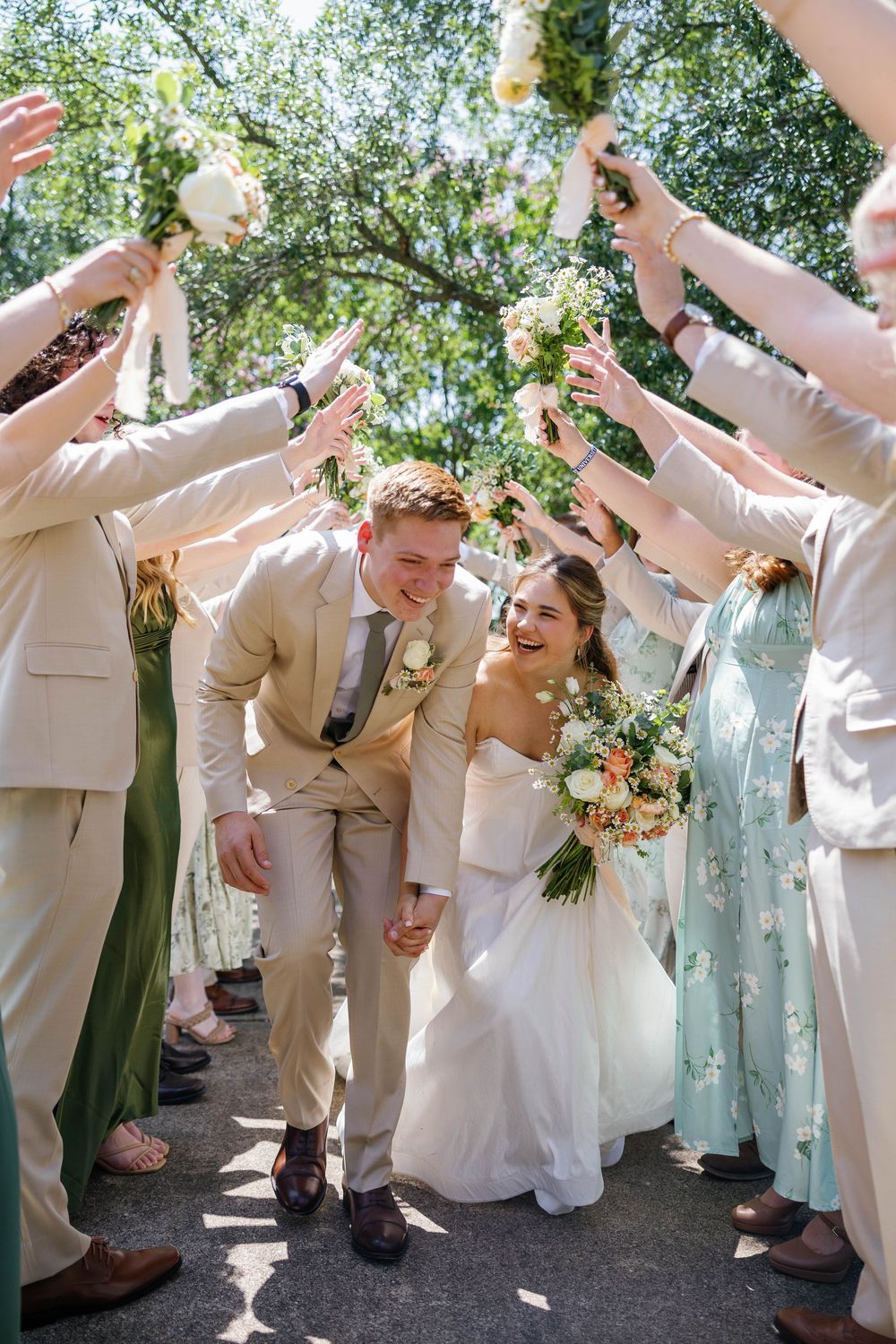 Joyful newlyweds walking through tunnel of raised bouquets held by wedding party outdoors.