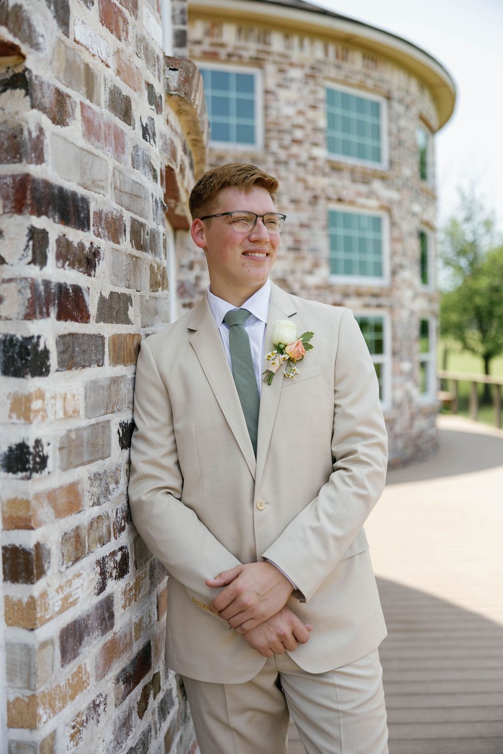 Smiling groom in a tan suit with floral boutonniere leaning against a brick wall outdoors.