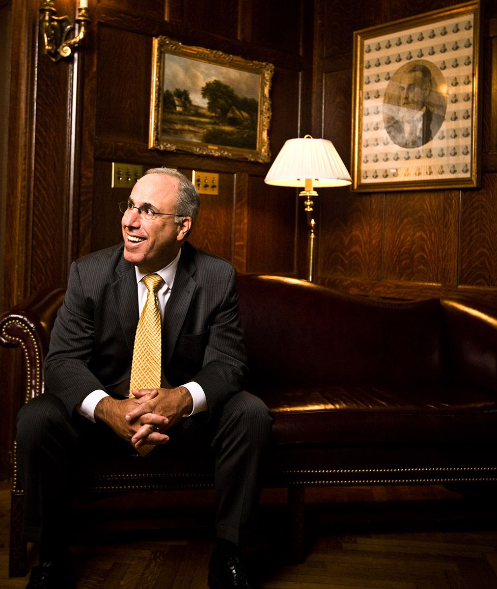 Smiling businessman in suit sits on leather couch in wood-paneled room with portrait on wall.
