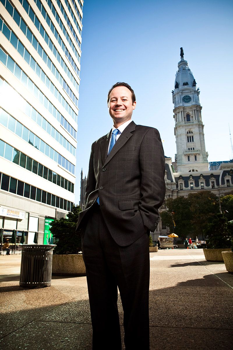Smiling businessman in dark suit stands in front of Philadelphia City Hall on a sunny day.