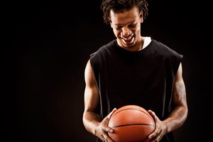 Young male basketball player in black sleeveless shirt holds basketball against dark background.