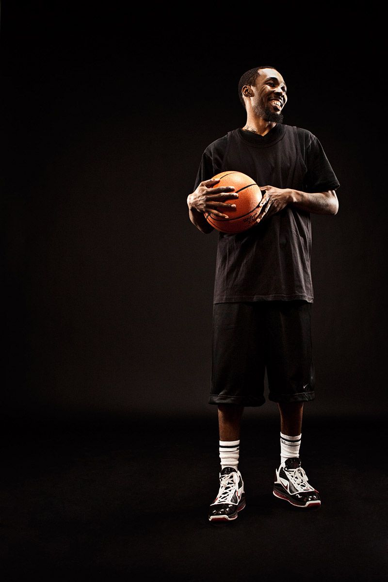 Smiling basketball player in black outfit holds orange basketball against dark background.