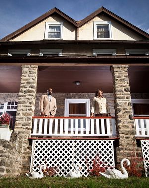 A couple stands on the porch of a stone house with white lattice railings and swan decorations.