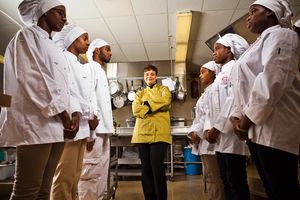 A chef in yellow instructs a group of culinary students in white uniforms in a professional kitchen.