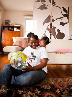 A smiling mother holds a globe while her young daughter hugs her from behind indoors.
