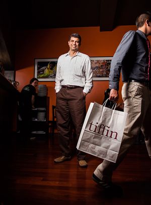 A smiling man stands in an office as another person carries a Tiffin branded shopping bag.