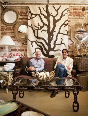 Two men sit on a rustic sofa in an eclectic antique shop with brick walls and decor.