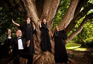 Four performers in black formal wear pose dramatically around a large tree outdoors.