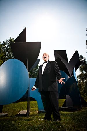 A man in a tuxedo sings passionately outdoors near large colorful abstract sculptures.