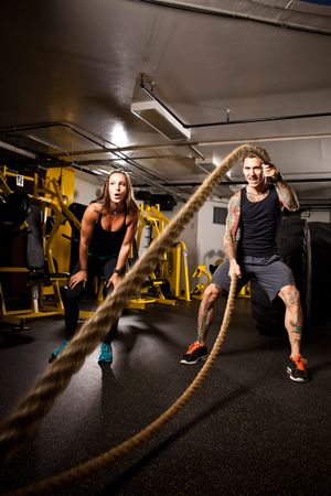 Two athletes exercise with heavy battle ropes in a dimly lit gym.