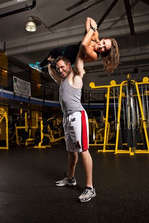 A male trainer lifts a smiling woman overhead in a well-equipped gym.